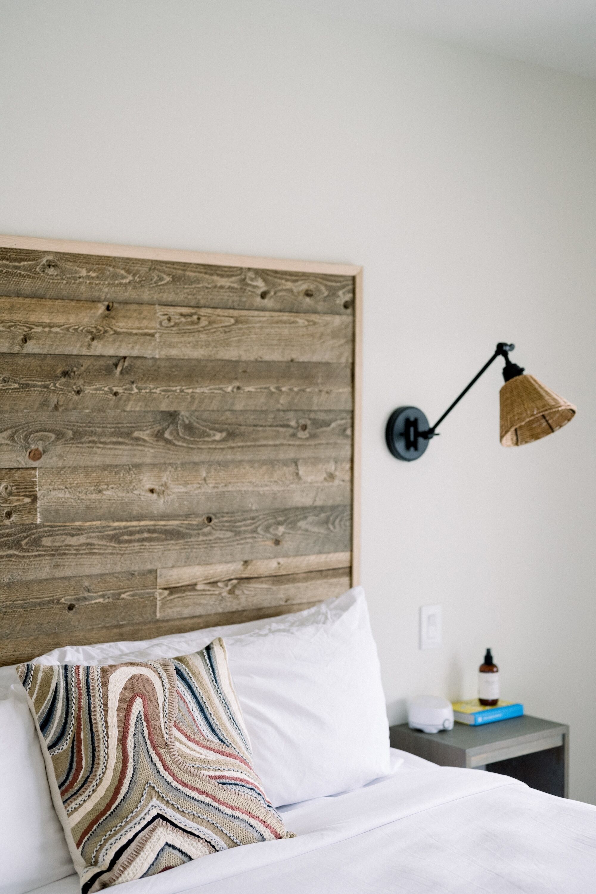 Ein Bett mit Holz-Kopfteil, einem gemusterten Kissen, einem Nachttisch mit Lampe und einer Wasserflasche.