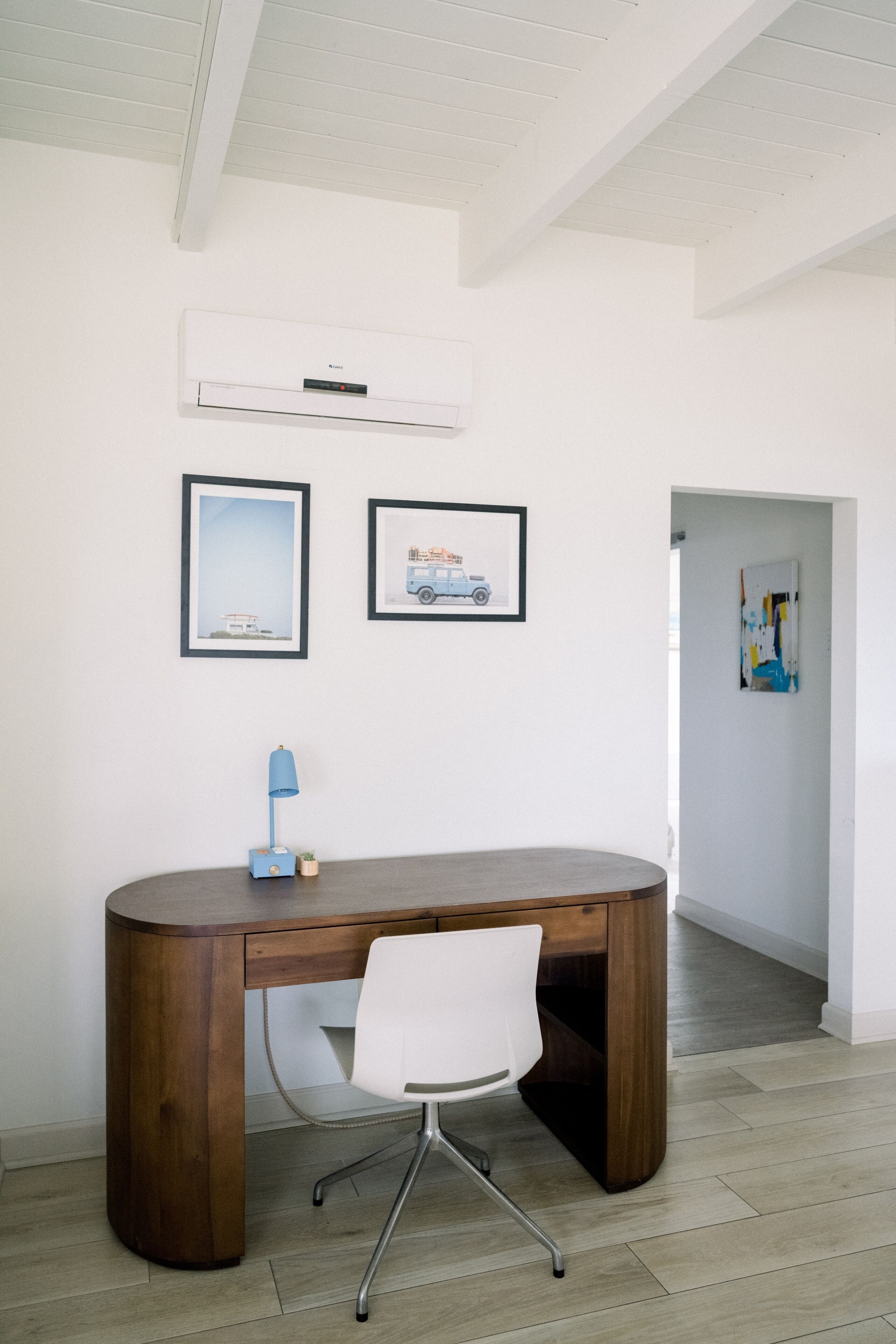 Un espacio de oficina moderno con un escritorio de madera, una silla blanca, cuadros enmarcados y un equipo de aire acondicionado en la pared.