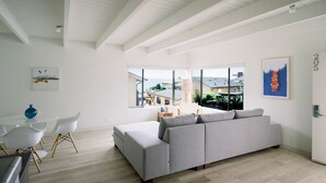 Un vaste espace de vie comprenant un grand canapé gris, des chaises blanches et une table à manger. De grandes fenêtres offrent une vue sur l’extérieur.