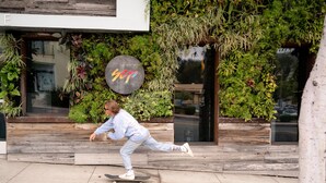 Une personne fait du skateboard sur un trottoir, devant un bâtiment à la façade en bois et orné d’un mur végétal.
