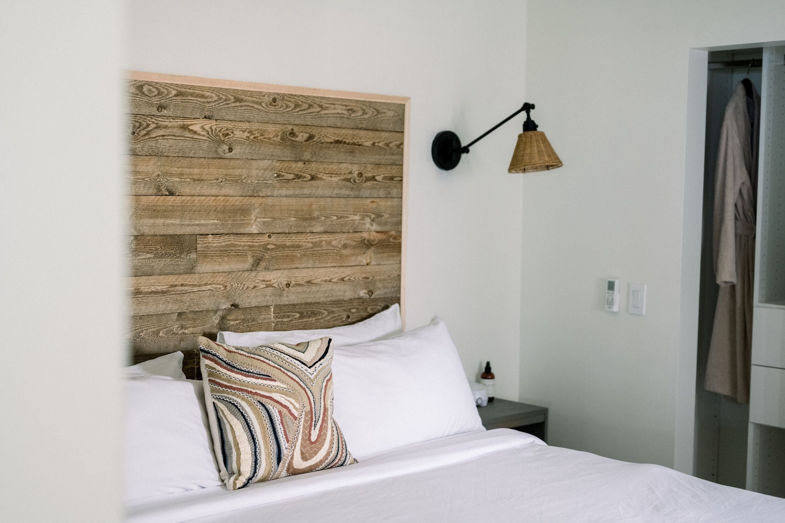 A bedroom with a wooden headboard, a bedside lamp, a bed with white linens, and a hanging robe.