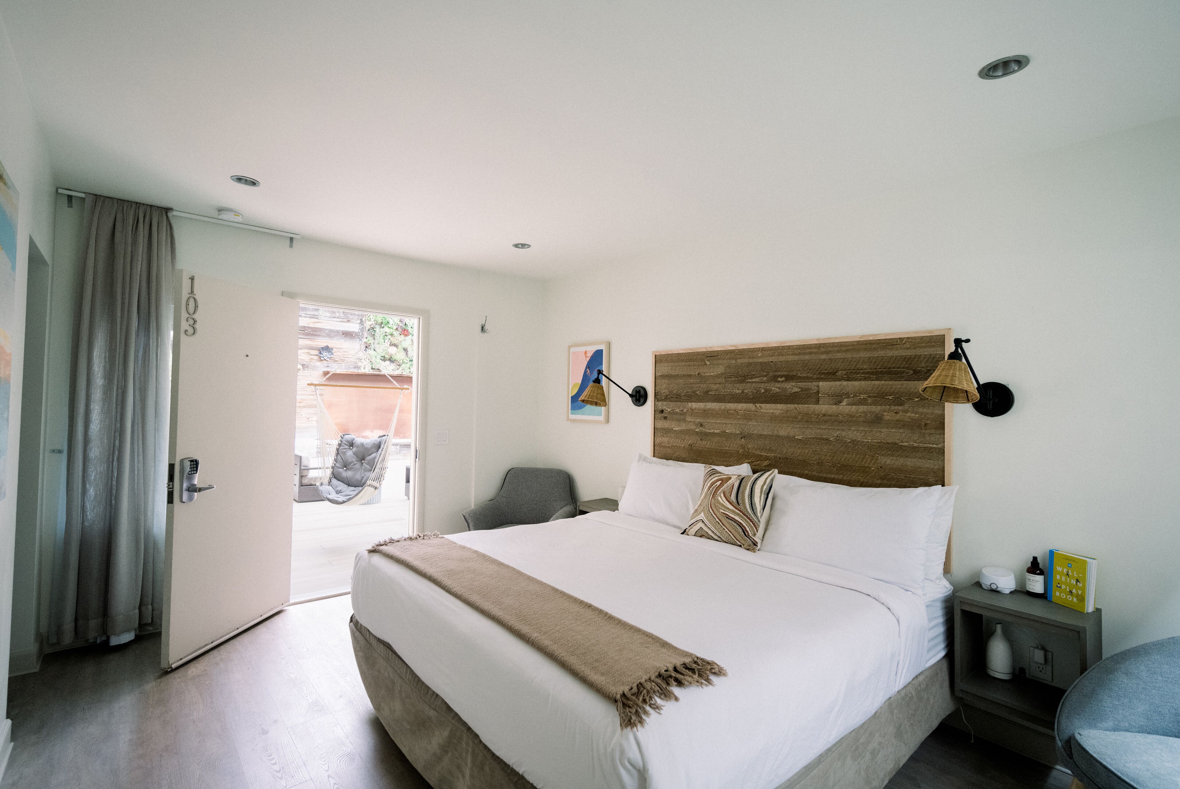 Habitación de hotel con una cama grande, cabecera de madera, mesita de noche con lámpara, una silla y una puerta que da a un espacio exterior.