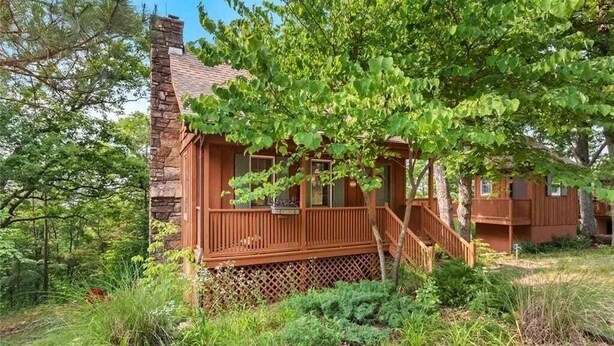 Une cabane rustique avec une cheminée en pierre, une terrasse en bois et un grand arbre verdoyant devant.