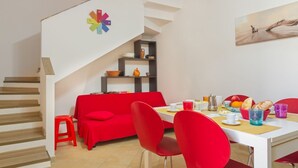 Ein moderner Essbereich mit einem roten Sofa, einem weißen Tisch und roten Stühlen. Im Hintergrund ist eine Treppe mit einer bunten Uhr zu sehen.