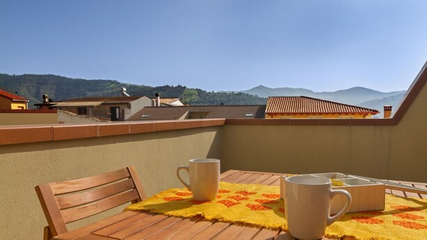 Ein Holztisch mit zwei weißen Tassen und einer gelben Tischmatte auf einem Balkon mit Blick auf Häuser und Hügel.