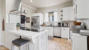 A modern kitchen with white cabinetry, a stainless steel refrigerator, a built-in oven, and a central island with a bar stool. - The Pendleton Lodge-8 bedrooms/3baths-heart of COS (Colorado Springs)