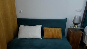 Un canapé de couleur turquoise agrémenté d’un coussin blanc et d’un coussin à rayures, une table en verre avec une lampe, et une petite table en bois.
