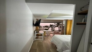 Une chambre avec un lit, un bureau, une chaise et un téléviseur fixé au mur. La fenêtre offre une vue sur des palmiers et un coucher de soleil.