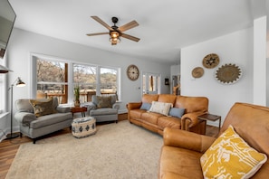 A living room with a ceiling fan, a large window, and a brown leather sofa.