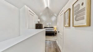 A narrow hallway with white walls, a wooden floor, and a sloped ceiling. There is a bed with a gray bedspread and a lamp on a bedside table. Framed artwork is on the walls. - Lansing 3BR near downtown — Game room & EV garage (Lansing)