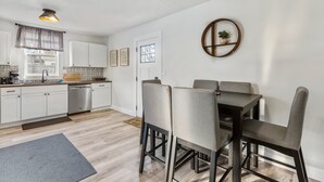 A modern kitchen with white cabinets, a dishwasher, and a dining area with a dark table and grey chairs. - Lansing 3BR near downtown — Game room & EV garage (Lansing)