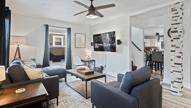 A modern living room with a ceiling fan, a fireplace, a TV mounted on the wall, and a stone pillar. - Lansing 3BR near downtown — Game room & EV garage (Lansing)