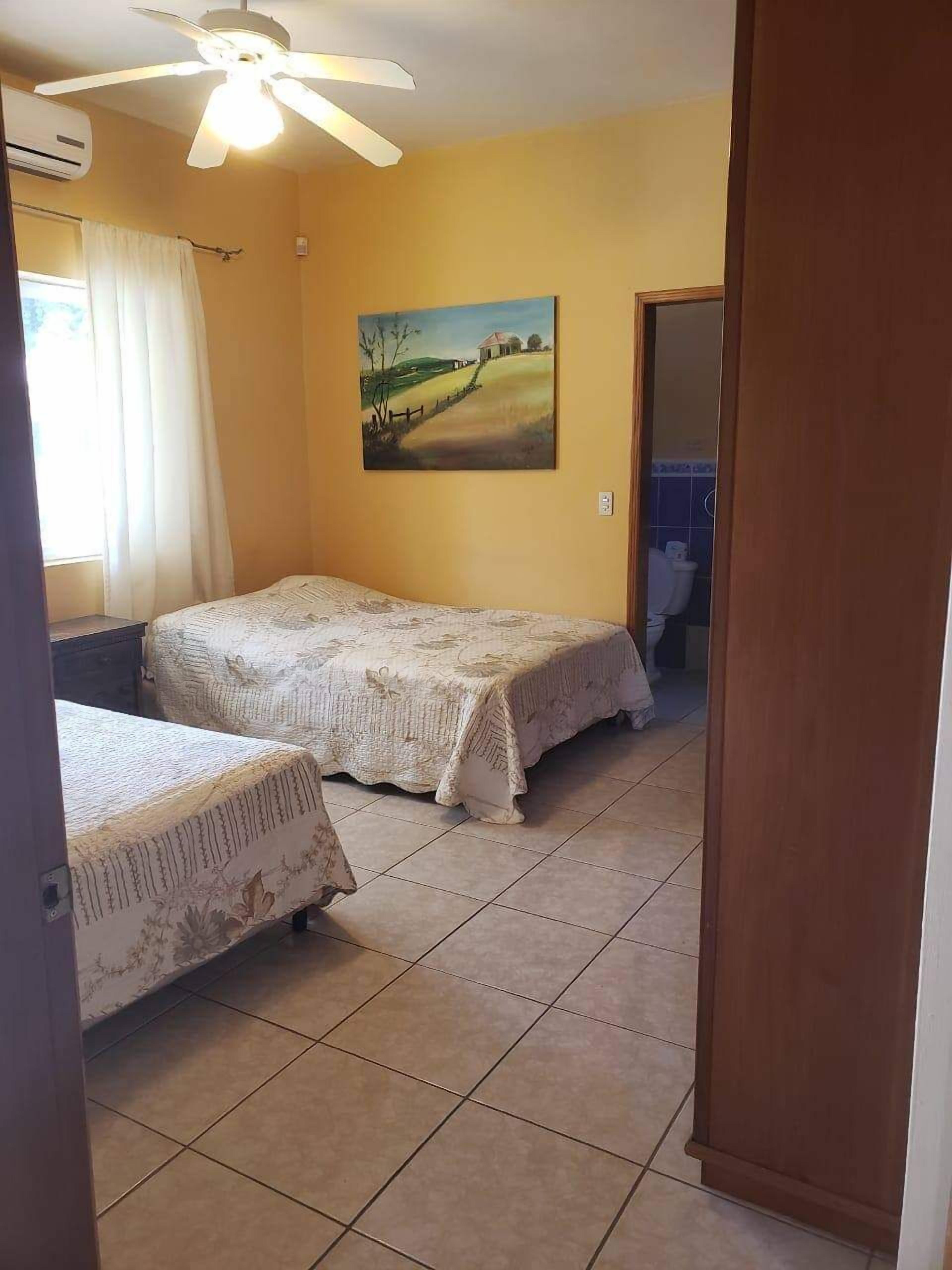 A bedroom with two beds, a ceiling fan, a painting on the wall, and a bathroom visible through an open door.