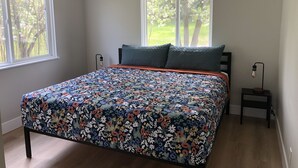 Une chambre à coucher avec une couvre-lit à motifs floraux, deux fenêtres, une table de chevet avec une lampe et une vue sur les arbres.
