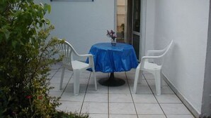 Un pequeño patio con una mesa redonda y dos sillas blancas, un mantel azul y una pared blanca con una franja azul.