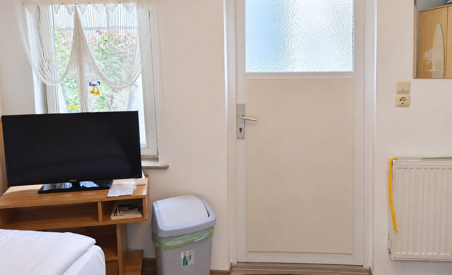 Une pièce comprenant un lit, un bureau avec un écran d’ordinateur, une poubelle, une porte et une fenêtre avec des rideaux.