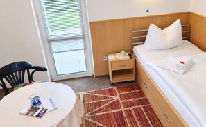 Una habitación de hotel con una cama, una mesita de noche, una mesa con una revista y una ventana con persianas.