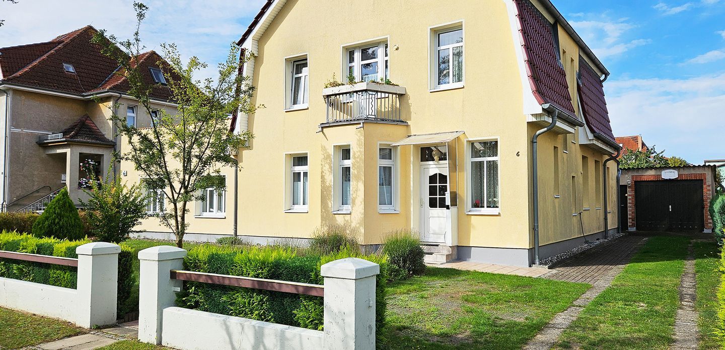 Ein zweigeschossiges Wohngebäude mit gelber Fassade, einem Balkon mit Metallgeländer und einem Ziegelportal.