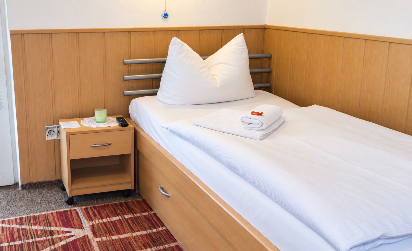 A single bed with a wooden headboard, a nightstand with a lamp, and a red patterned rug.