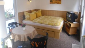 Una habitación pequeña y funcional con una cama, televisión, una mesa y sillas, y un ventanal.