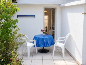 A small patio with a round table covered by a blue tablecloth, two white plastic chairs, and a vase with pink flowers. - Möwe 1 - Holiday apartment Möwe 1 with terrace near the sandy beach in Kühlungsborn (Kühlungsborn)