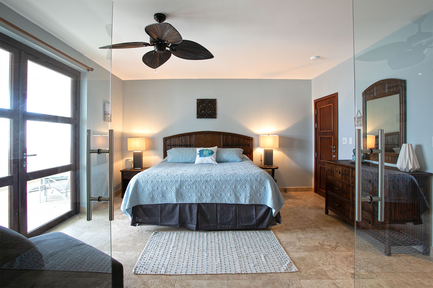 A bedroom with a large bed, two bedside tables with lamps, a ceiling fan, a large mirror, and a sliding glass door leading to a balcony.
