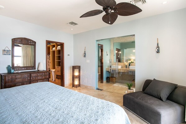 A bedroom with a large bed, a dark sofa, a wooden wardrobe, a ceiling fan, and a bathroom visible through a glass door.