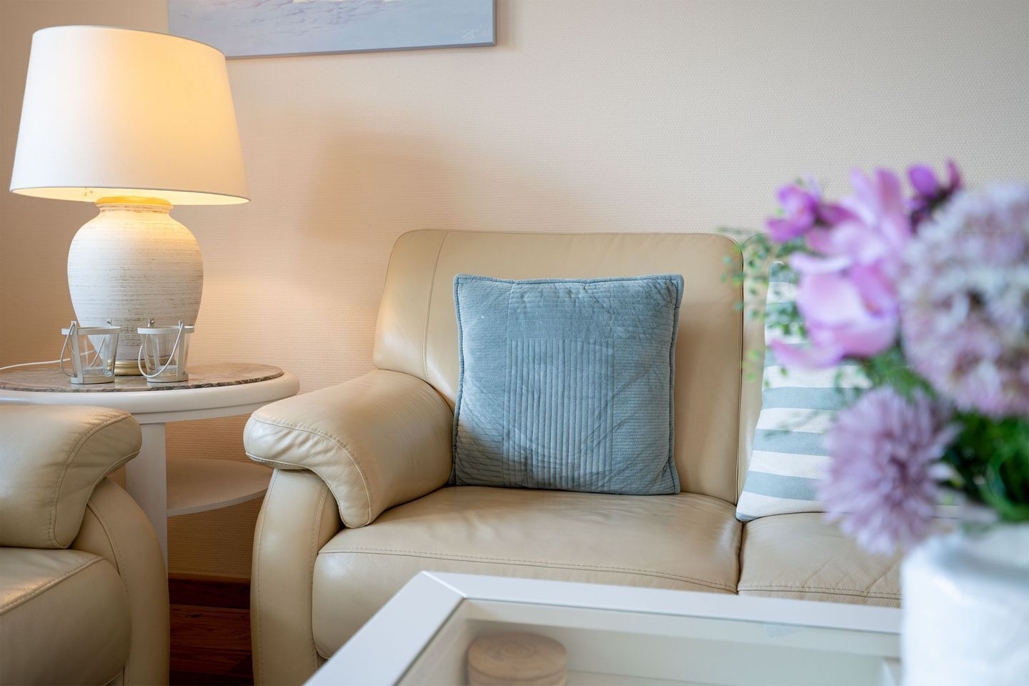 Een beige leren fauteuil met een blauw kussen, een witte bijzettafel met een lamp en een vaas met bloemen.