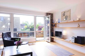 A modern living room with a glass door leading to a balcony, a flat-screen TV, a sound system, and a bookshelf.
