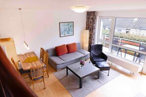 A modern living room with a grey sofa, a black leather chair, a wooden dining table, and a coffee table. There is a painting on the wall and a window with a view of buildings. - (BEE1) Apartment Ernstorfer - Apartment "Ernstorfer" for 3 persons ( (Timmendorfer Strand)