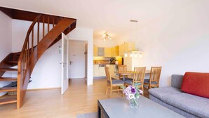Un appartement moderne avec un escalier en bois, un coin repas avec des chaises en bois, un canapé gris et une petite table sur laquelle est posé un vase de fleurs.