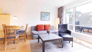 Un salon moderne avec un canapé gris, deux chaises en bois, un fauteuil en cuir noir, une table basse et une grande fenêtre donnant sur l’extérieur.