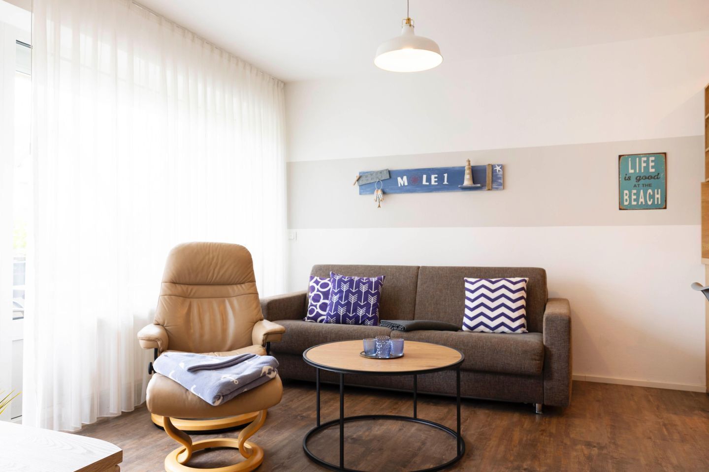 Ein gemütliches Wohnzimmer mit einem Ledersessel, einem grauen Sofa und einem runden Couchtisch. Dekorative Kissen und ein Wandschild mit der Aufschrift 
