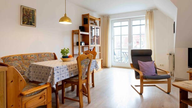 A cozy living room with a wooden sofa, a dining table with a floral tablecloth, a rocking chair, and a bookshelf.