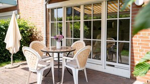 A patio with a round table and four chairs, a white umbrella, and a large window with a view of greenery. - (OST1) Holiday apartment "Lörper 1" - Holiday apartment "Lörper 1" up to 3 persons (Timmendorfer Strand)