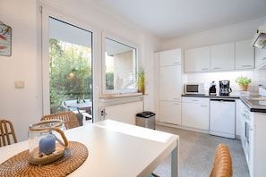 A modern kitchen with white cabinets, a dining table with chairs, a window overlooking a garden, and a microwave. - (MEI71) Apartment "Strandlust" - Apartment "Strandlust" up to 4 persons (Timmendorfer Strand)