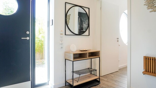 A modern hallway with a dark door, a round window, a mirror, a small wooden console table, and a radiator.