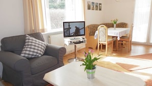 Uma sala de estar com um sofá cinzento, uma mesa pequena, uma televisão, uma mesa de jantar com cadeiras e uma janela com cortinas.