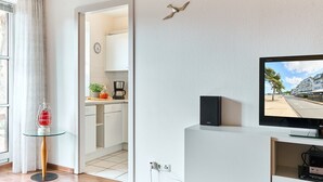 A modern living room with a flat-screen TV, a glass table with a red lantern, a kitchen visible through an open door, and a wall-mounted bird decoration. - (OST35c) Holiday apartment "Sonnendeck" - Holiday apartment "Sonnendeck" for 2 persons (Timmendorfer Strand)