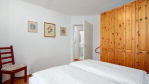 Un dormitorio con un armario de madera, una cama con ropa de cama blanca y una silla con cojín floral.