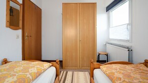 A bedroom with two beds, a wooden wardrobe, a small stool, and a window with blinds.