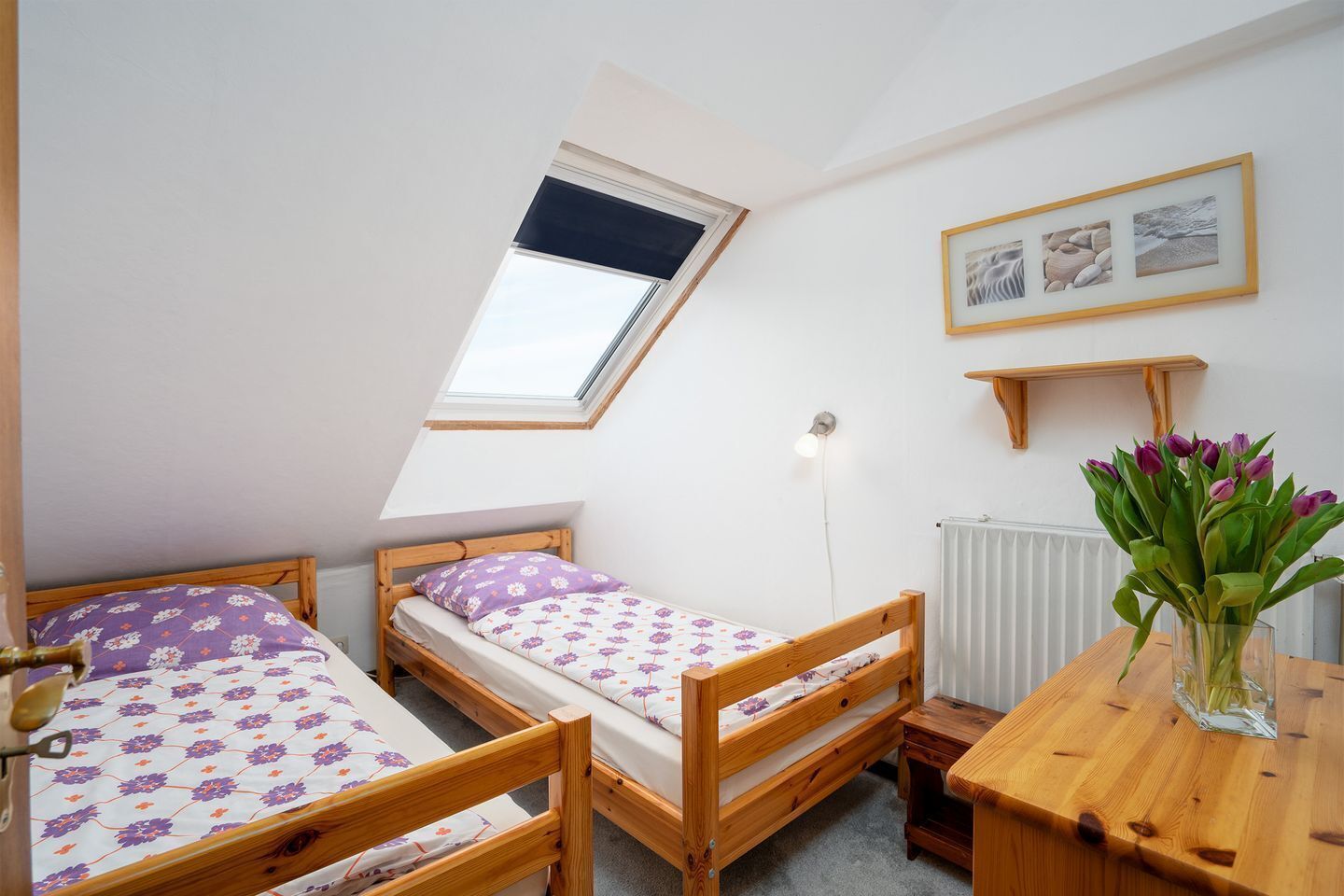 A small, cozy room with two wooden beds, a skylight, a radiator, and a wooden table with a vase of flowers.