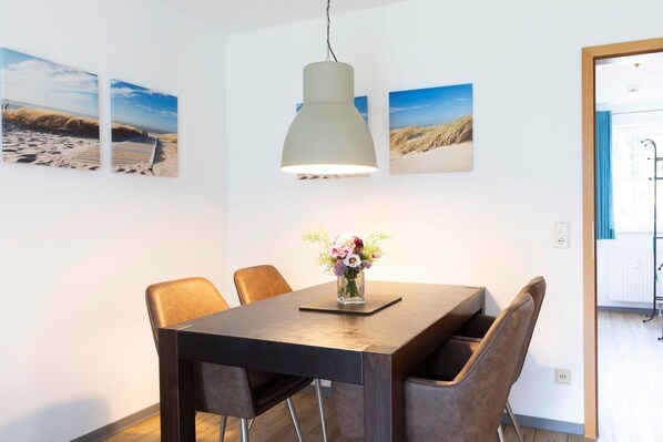 Comedor con una mesa de madera, seis sillas, una lámpara colgante y tres cuadros con motivos playeros.