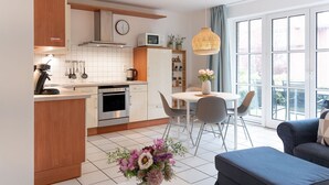 A modern kitchen with white cabinets, a stainless steel oven, and a microwave. There is a dining area with a white table and chairs, and a sofa in the living room. - (OST8) Holiday apartment "Lörper 2" - Holiday apartment "Lörper 2" up to 4 persons (Timmendorfer Strand)