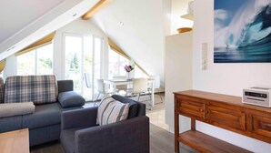 Ein modernes Wohnzimmer mit einer Couch, einem Couchtisch und einer Holzkommode. Es gibt eine Essecke mit Tisch und Stühlen, und ein großes Fenster lässt viel natürliches Licht herein.