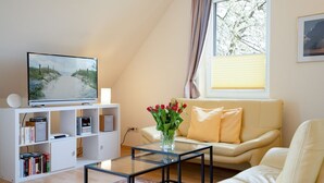 A modern living room with a television on a white entertainment unit, a glass coffee table, a beige sofa, and a vase with red flowers. - (AMW1) Apartment Garbers/Thiele - Apartment "Garbers/Thiele" to 3 (Timmendorfer Strand)