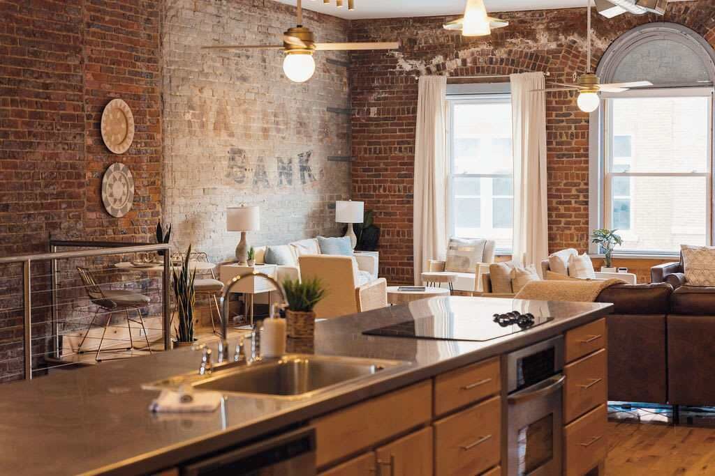 A kitchen with a large island, a dining area with a brick wall, and a living room with a sofa and a window.