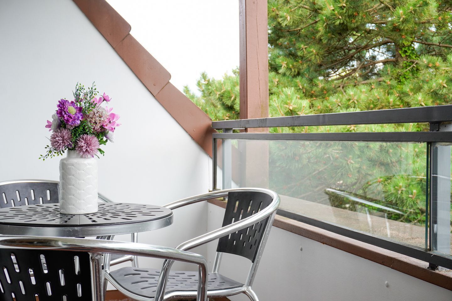 A balcony with a table and chairs, a white vase with flowers, and a view of trees through a window.