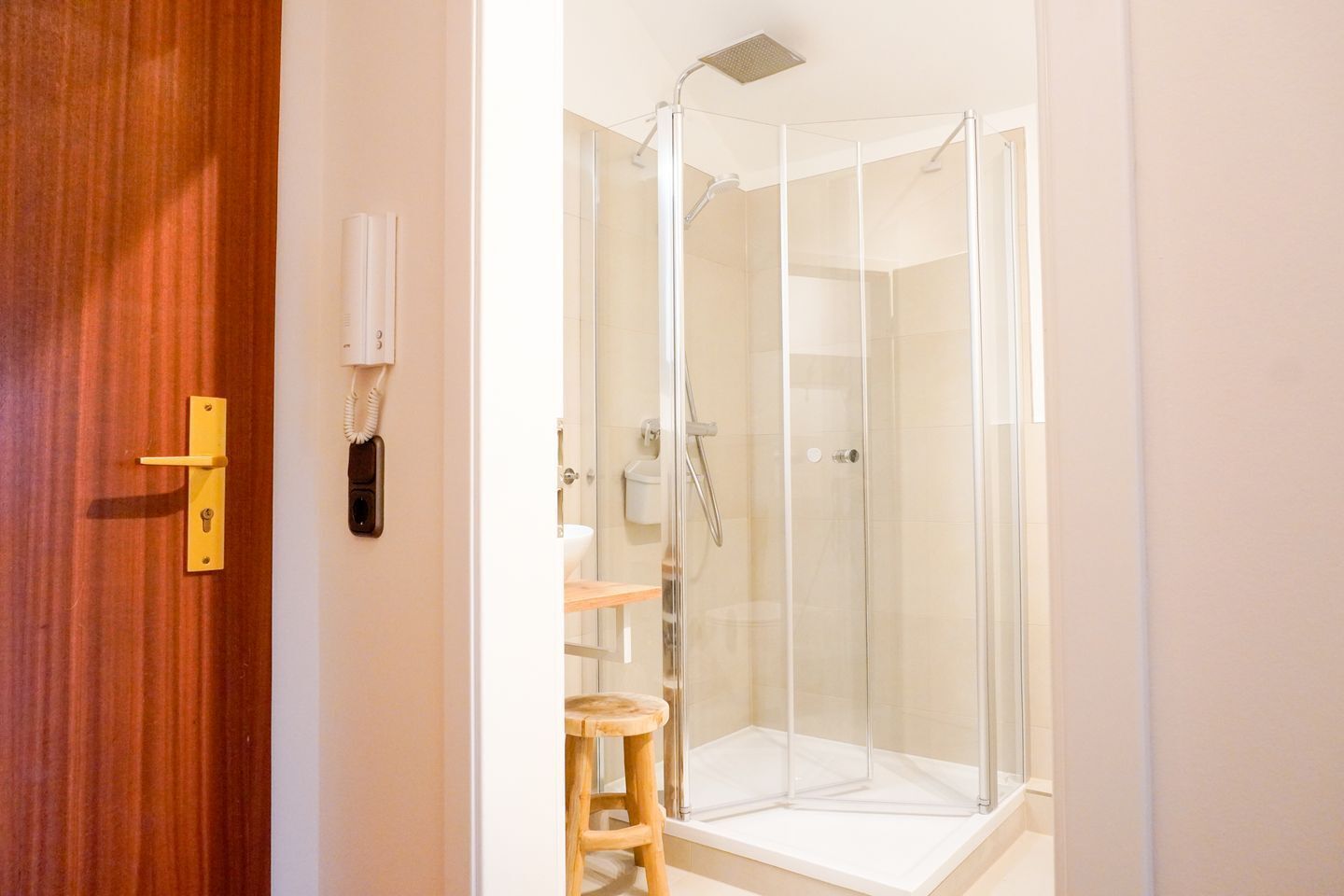 Une salle de bain avec une porte en bois, une douche fermée par des parois de verre et un tabouret en bois.