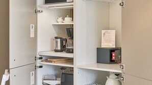 Una cocina equipada con microondas, hervidor, cafetera y máquina de café. Hay una estantería con un hervidor y una caja de cápsulas de café.